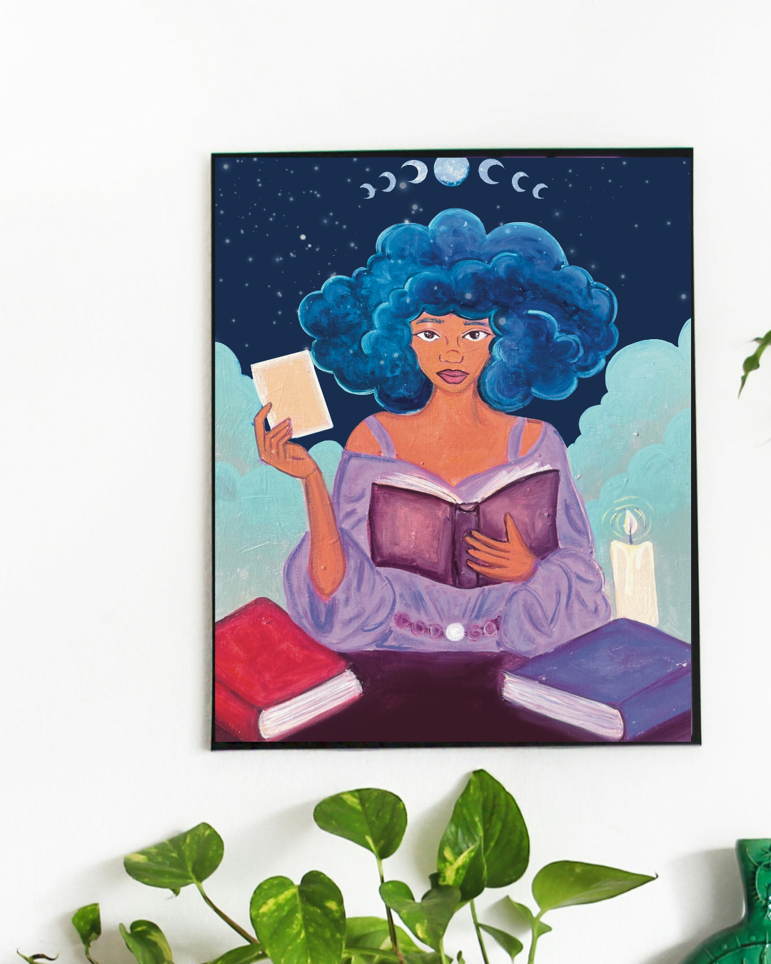 Framed artwork of a woman with blue hair holding a book and a tarot card, with 2 more books on the table and a candle. 